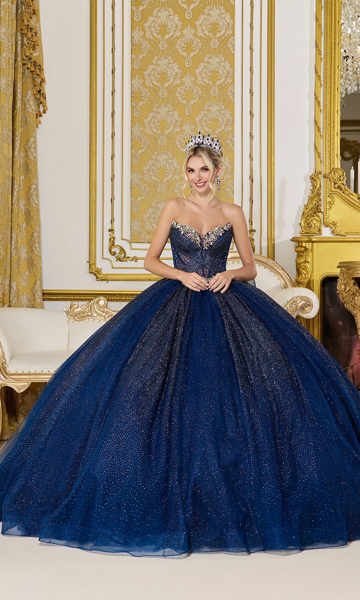 A woman in a navy Quinceanera Dress 5078 By Dancing Queen stands in an ornate, gold-trimmed room with classical decor and a white upholstered bench, wearing a tiara and a strapless corset bodice ball gown.